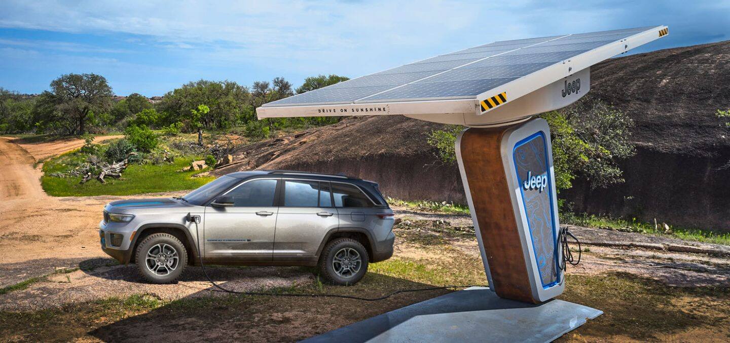 Display The 2022 Jeep Grand Cherokee 4xe Trailhawk parked on a dirt road beside a Jeep-branded solar-powered charging station.