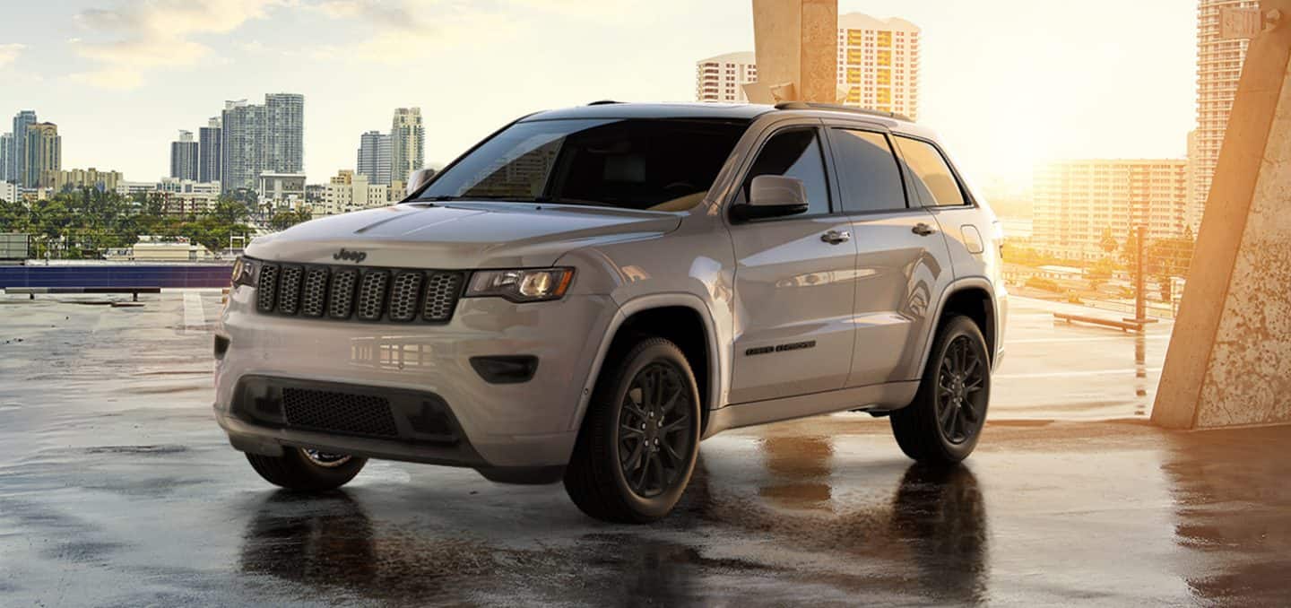 Display The 2022 Jeep Grand Cherokee WK Laredo X with the Altitude Appearance Package parked in an open square with a city skyline behind it and the sun setting in the distance.