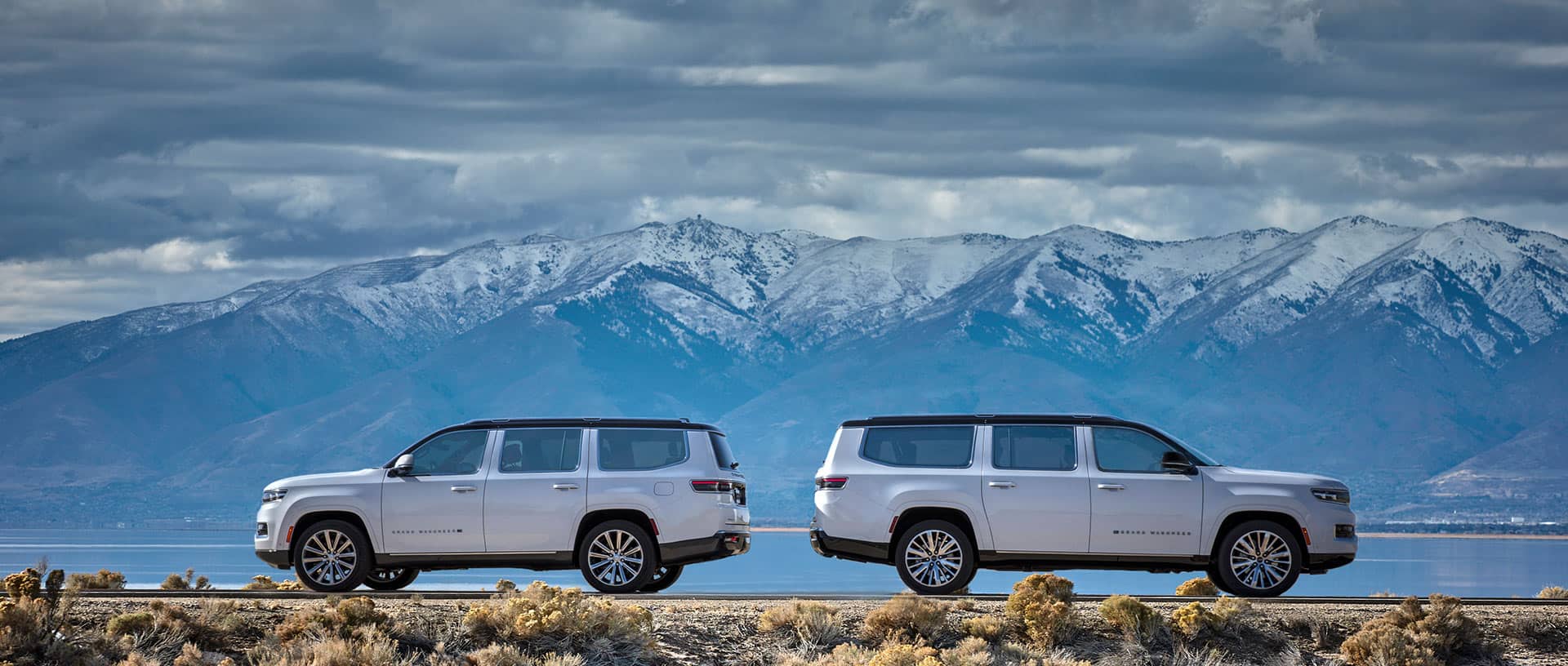 A 2023 Grand Wagoneer Series III Short Wheelbase and 2023 Grand Wagoneer Series III L Long Wheelbase parked back-to-back on a highway with snow-capped mountains in the background.