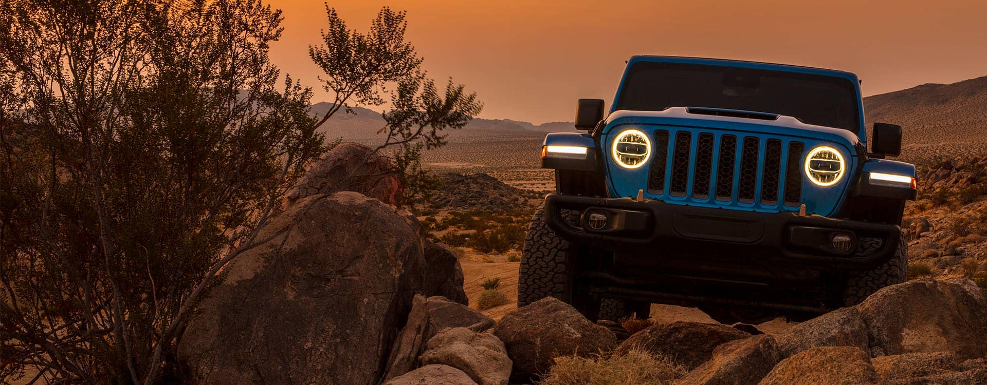 A head-on view of a 2022 Jeep Wrangler Rubicon 392 with its headlamps on, crawling over boulders with a bright orange sky at dusk in the background.