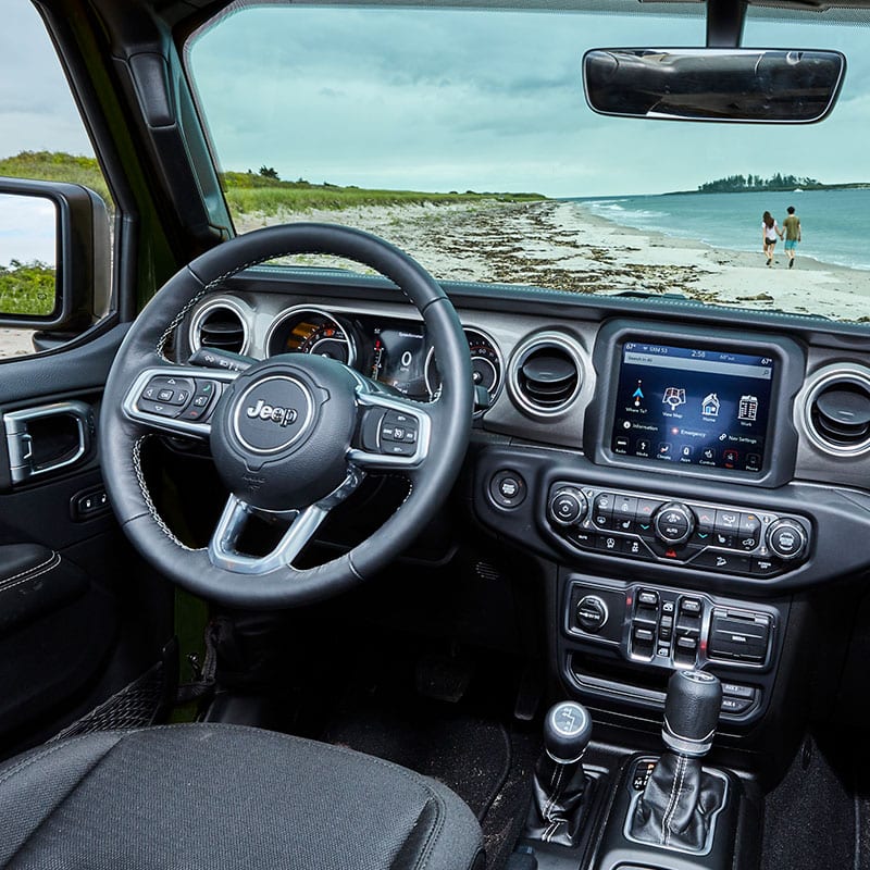 The steering wheel, Uconnect touchscreen and center stack controls on the 2022 Jeep Wrangler Sahara, parked at a beach.