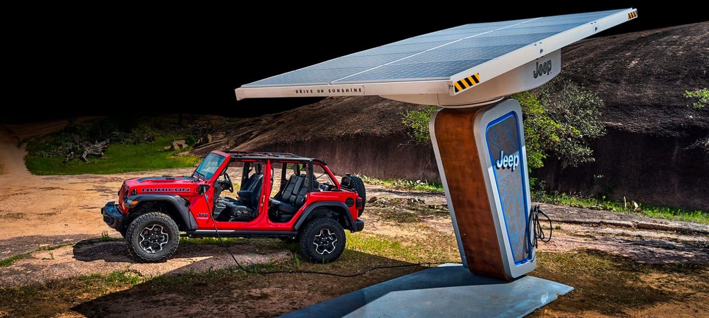 Un Jeep Wrangler Rubicon 4xe 2022 estacionado junto a una estación de carga de la marca Jeep en el bosque, mientras carga la batería de su motor eléctrico.