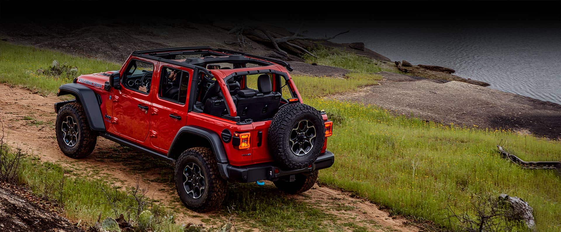 A Jeep Wrangler Rubicon 4xe with its roof off as it is driven on a dirt road beside a lake.