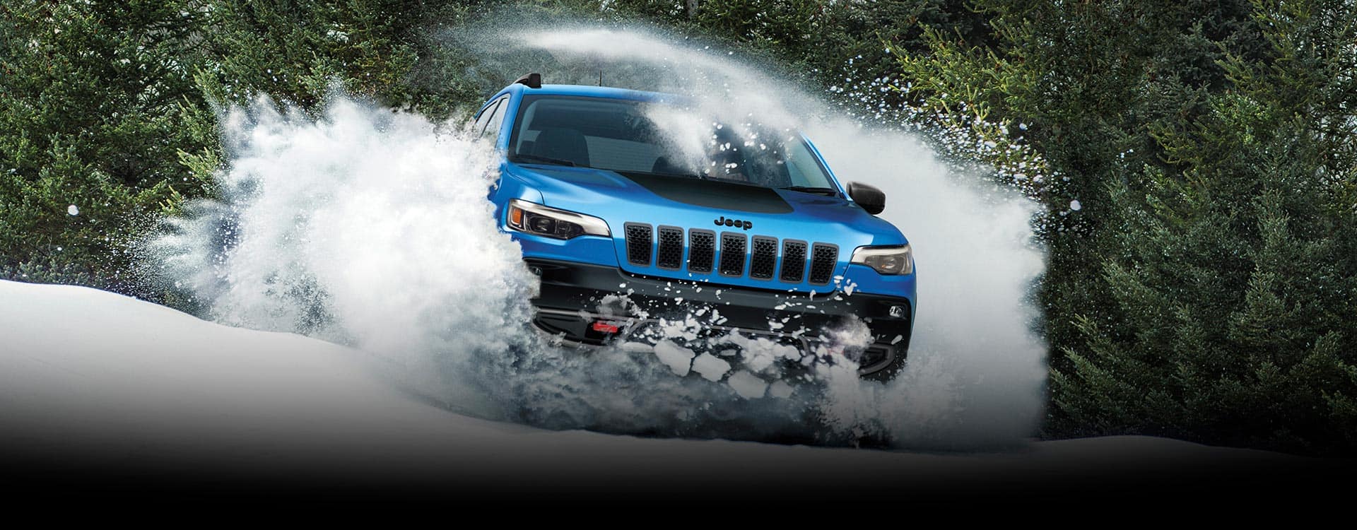 Un Jeep Cherokee Trailhawk 2023 atravesando un montículo de nieve, con una nube de polvo blanco emergiendo a ambos lados y en el techo del vehículo, con vegetación de hojas perennes de fondo.