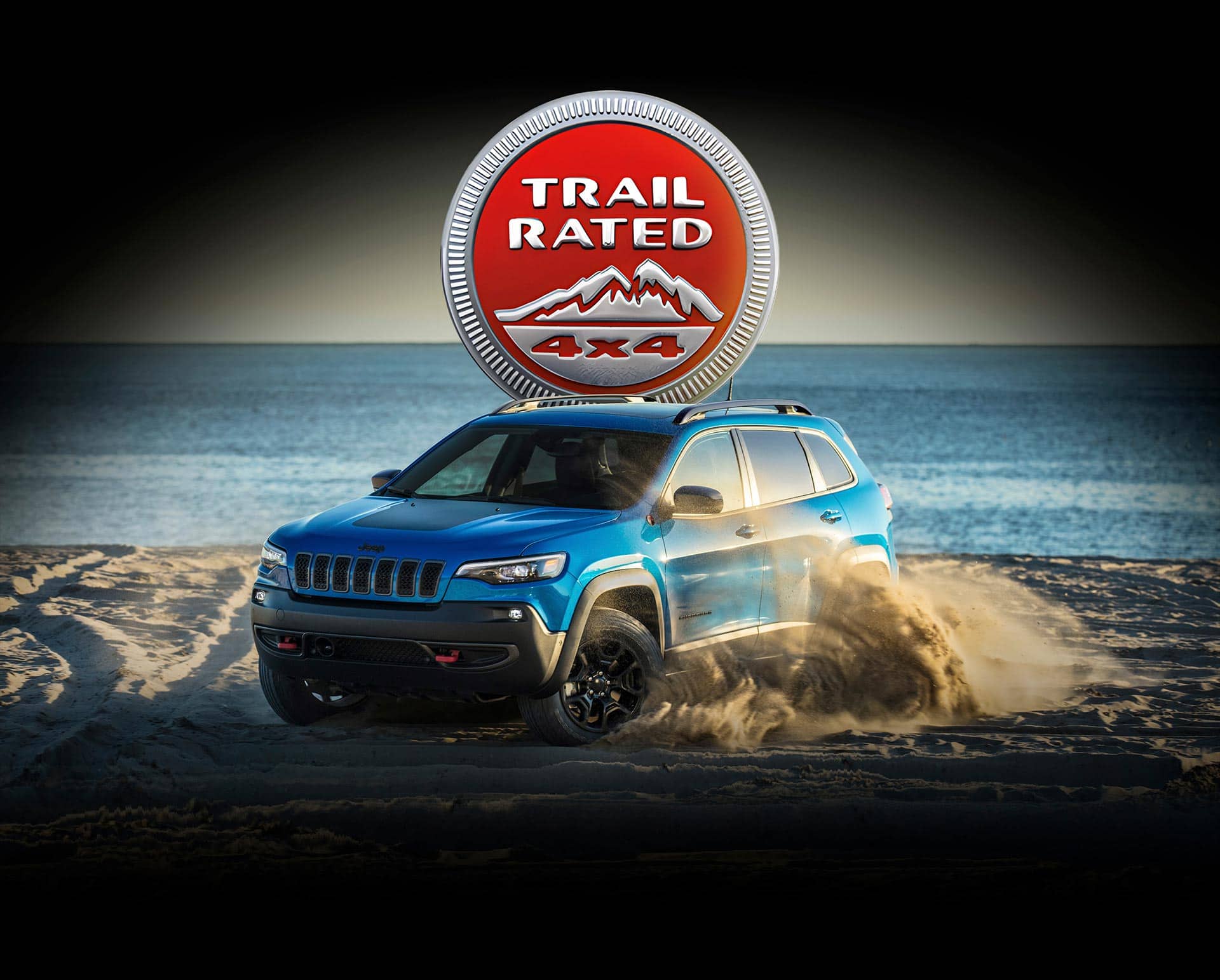 The Trail Ratted 4x4 badge. A blue 2023 Jeep Cherokee Trailhawk kicking up sand on a beach beside crystal blue water.
