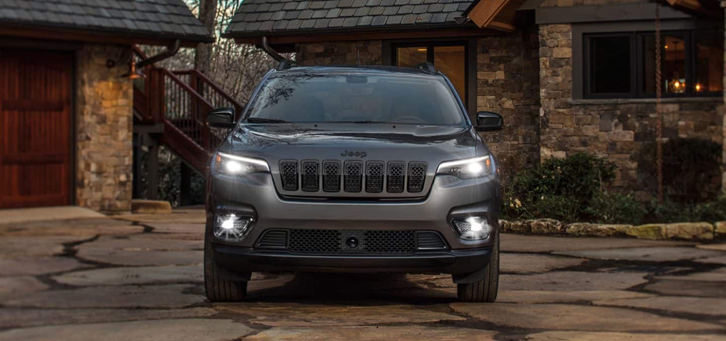 Display A head-on view of a 2023 Jeep Cherokee Altitude Lux with its headlamps and fog lamps on, parked in the driveway of a brick residential building.