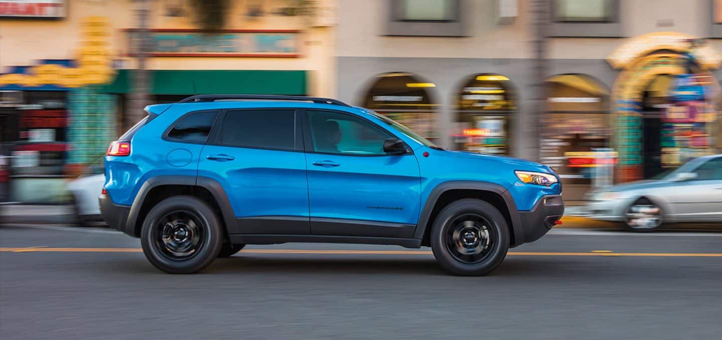 Display A profile view of a blue 2023 Jeep Cherokee Trailhawk being driven on a city street with the background blurred, indicating the vehicle is in motion.