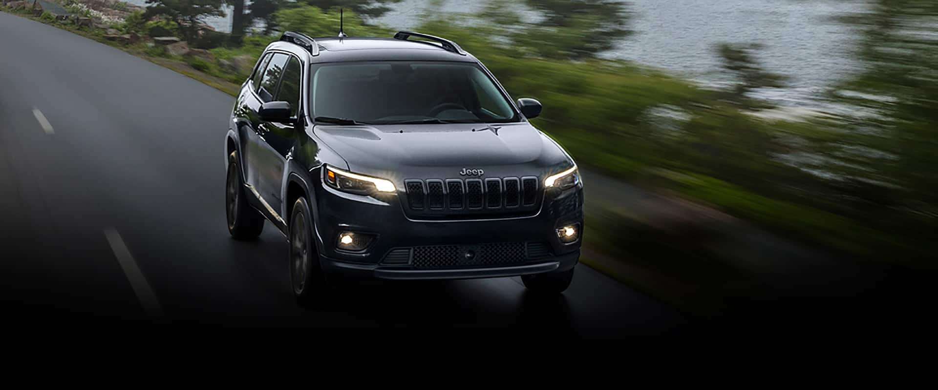 Un Jeep Cherokee Altitude Lux 2023 circulando por la carretera junto a un lago, con el cielo nublado y el fondo difuminado para indicar que el vehículo está en movimiento.