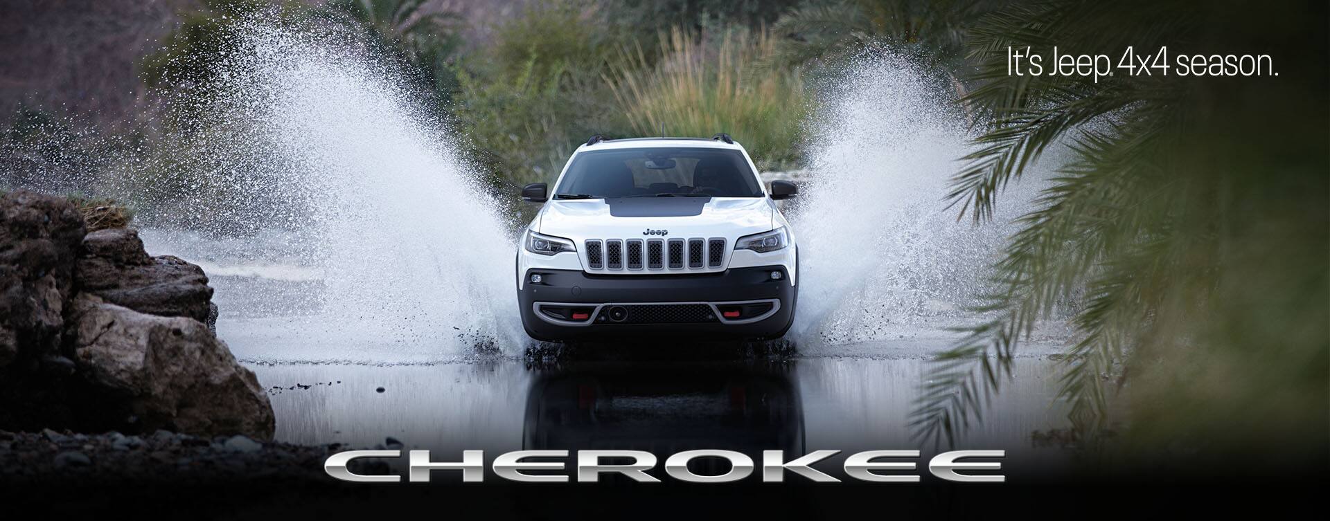 A head-on angle of a white 2023 Jeep Cherokee Trailhawk fording a stream off-road, with water splashing well above the top of the vehicle. Cherokee. The It's Jeep Season logo.
