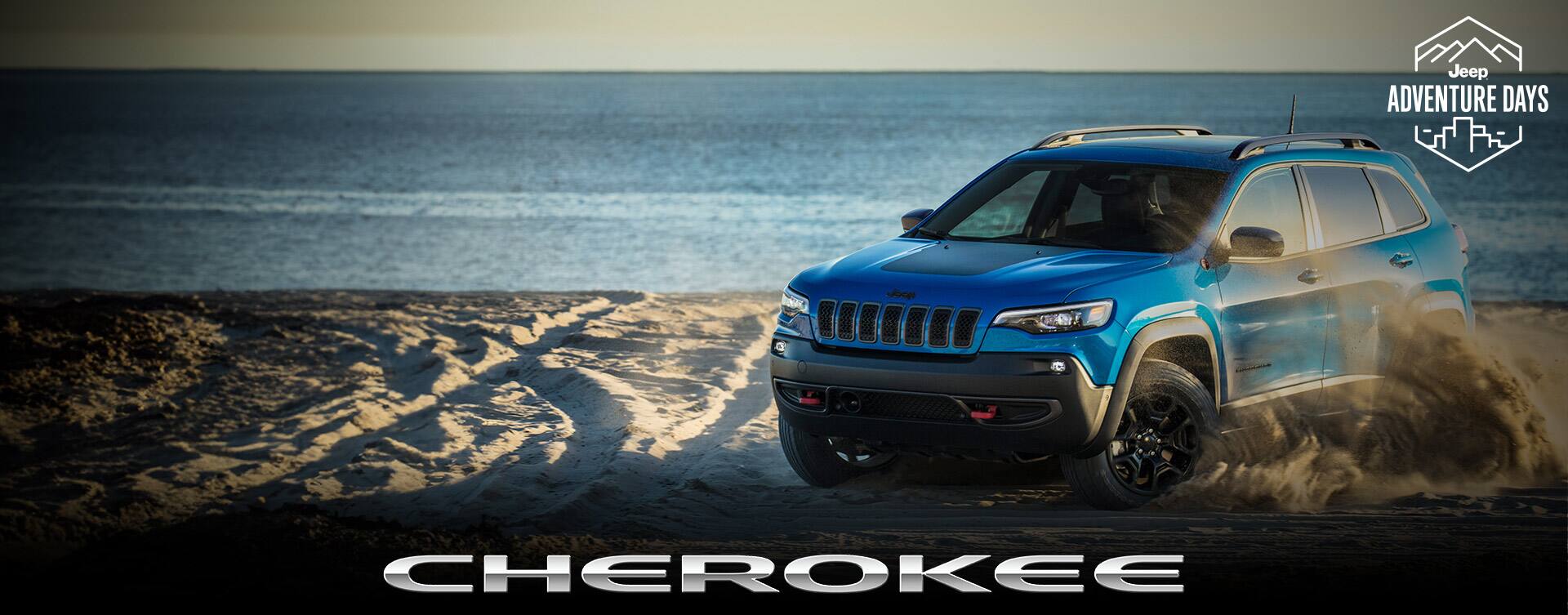 A blue 2023 Jeep Cherokee Trailhawk being driven on a beach, kicking up swirls of sand as it moves. Jeep Adventure Days logo. Cherokee.