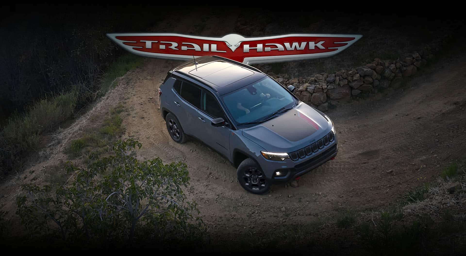 A 2023 Jeep Compass Trailhawk being driven on a dirt road hill beside a stone fence. The Trailhawk badge.