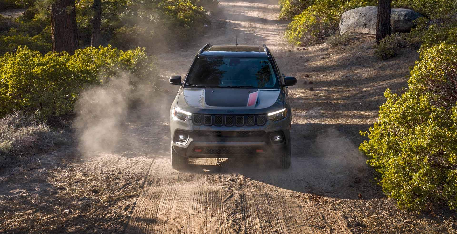 A raised head-on view of a 2023 Jeep Compass Trailhawk being driven off-road through a wooded trail.