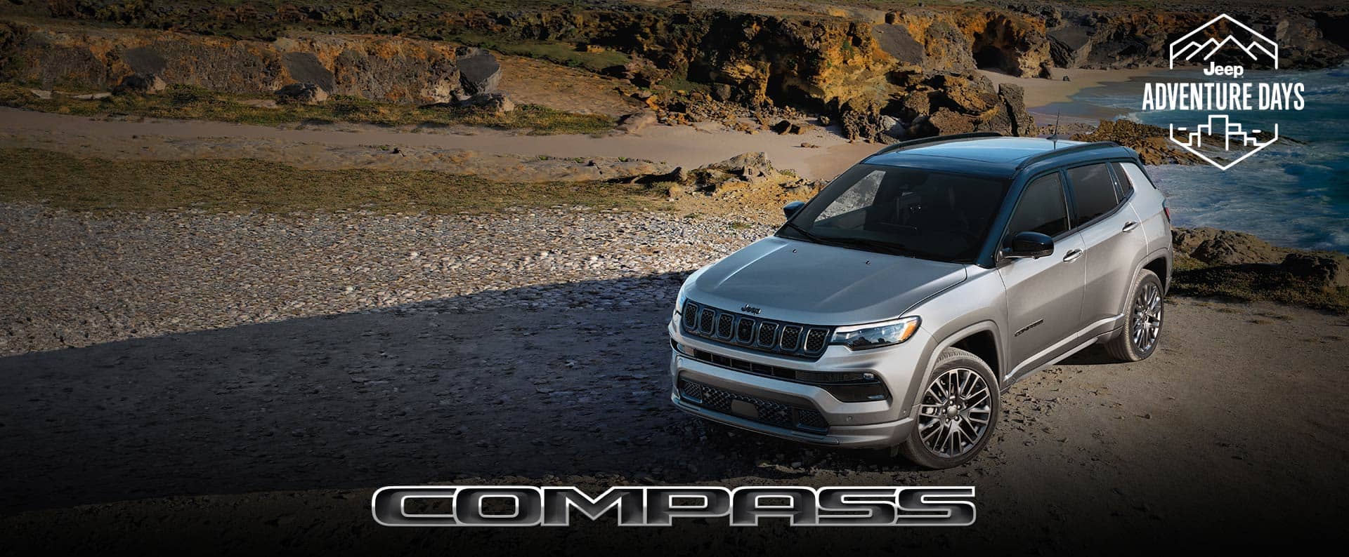 A driver-side front profile of a silver 2023 Jeep Compass High Altitude parked on a beach. Jeep Adventure Days logo. Compass.