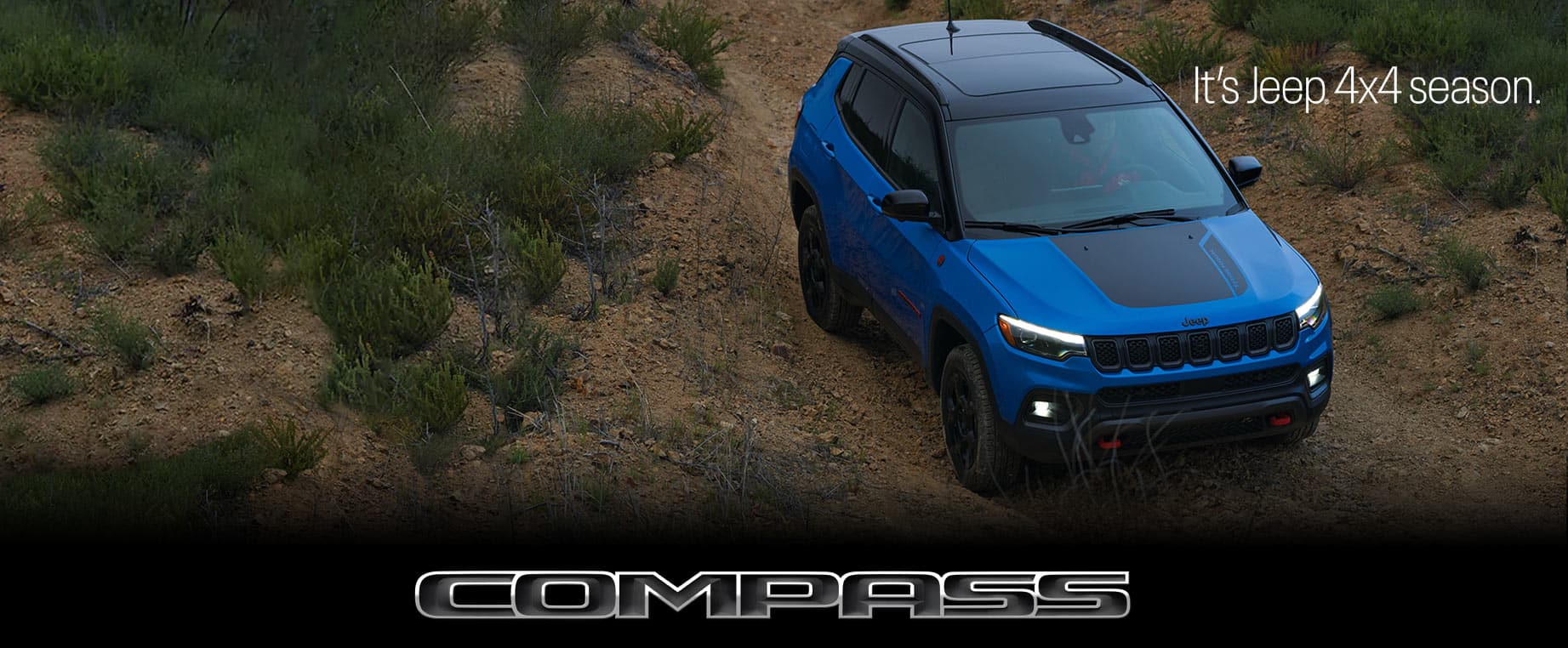 A raised angle of a blue 2023 Jeep Compass Trailhawk parked off-road. Compass. The It's Jeep Season logo.
