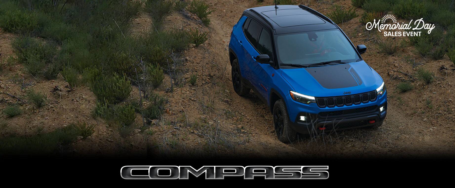 Ángulo elevado de un Jeep Compass Trailhawk 2023 azul estacionado fuera de la carretera. Compass. Logo del evento de ventas por el Día de los Caídos.