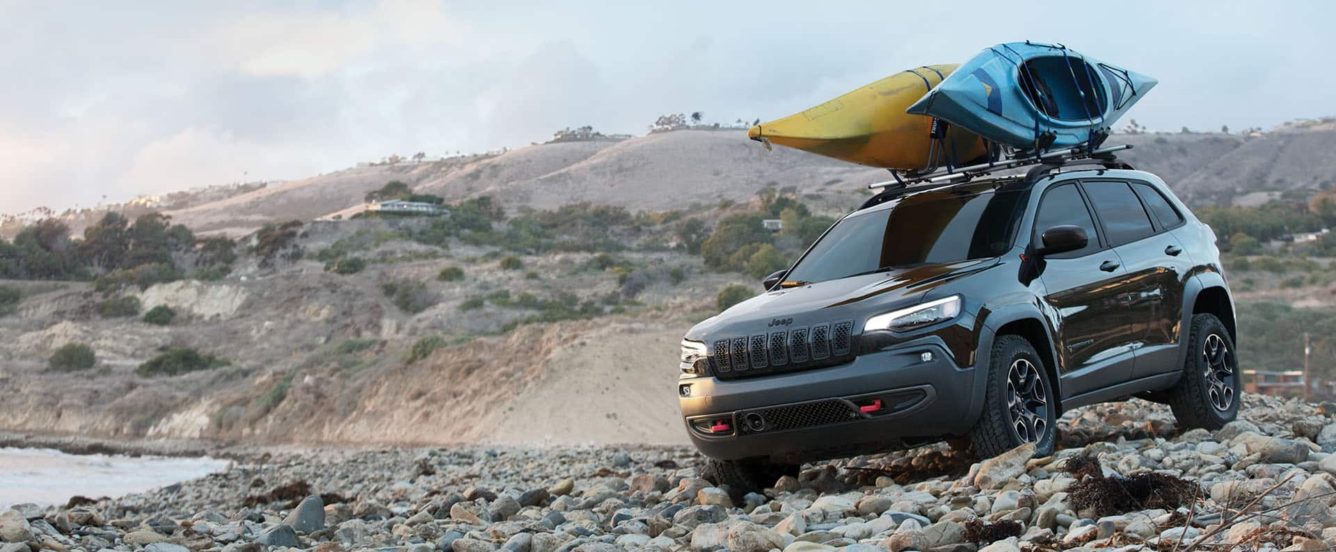 Un Jeep Cherokee Trailhawk 2023 con dos kayaks atados a su parrilla portaequipaje, avanzando sobre las piedras de una playa rocosa.