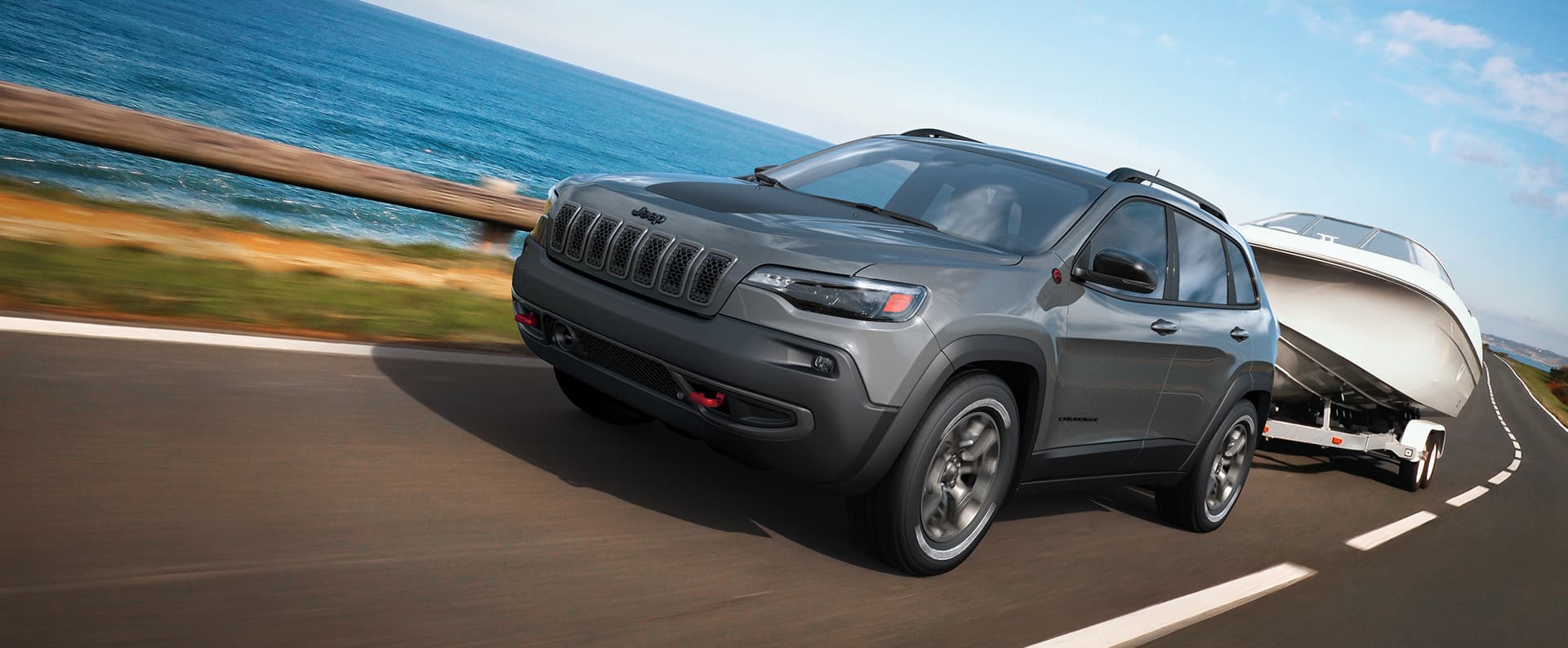 A 2023 Jeep Cherokee Trailhawk towing a motorboat, as it's driven on a highway beside a large body of water.