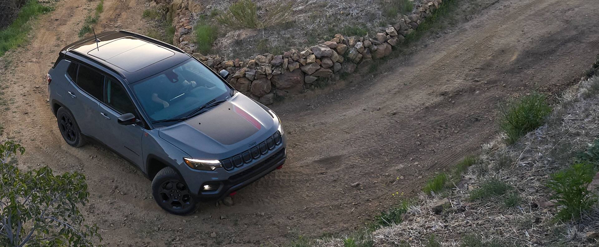 A 2023 Jeep Compass Trailhawk being driven on a dirt road hill beside a stone fence.