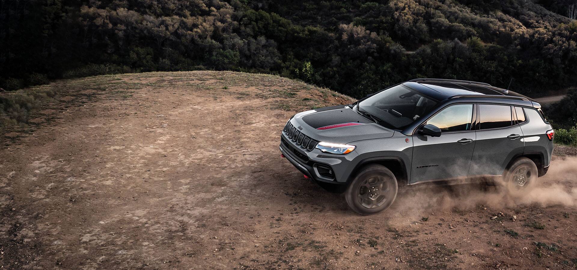 A 2023 Jeep Compass Trailhawk being driven off-road on a clearing, with a forest in the background.