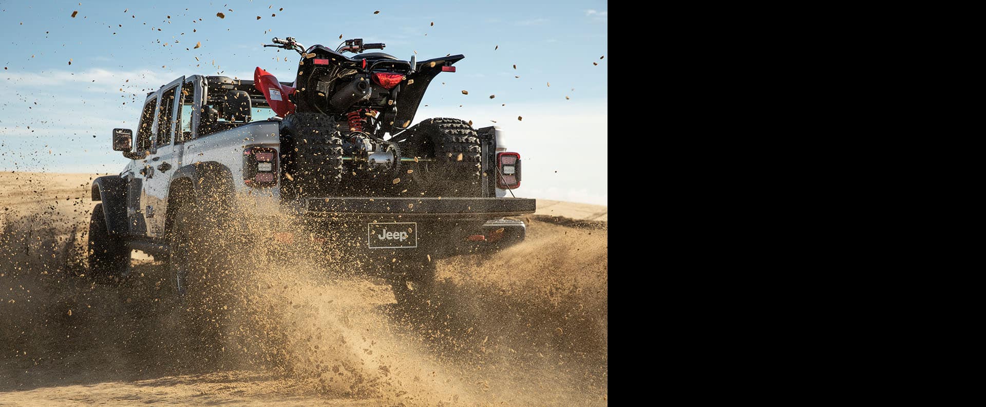 The 2023 Jeep Gladiator with an all-terrain vehicle in its truck bed, being driven on loose sand.