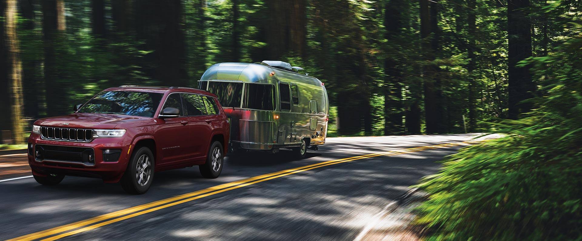 A red 2023 Jeep Grand Cherokee Overland being driven on a highway in the country, towing a travel trailer.