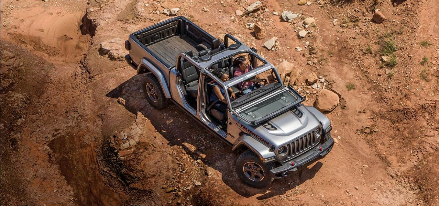 Display An overhead view of a silver 2023 Jeep Gladiator Rubicon with its doors and roof off and its windshield folded down, being driven down a dirt hill.