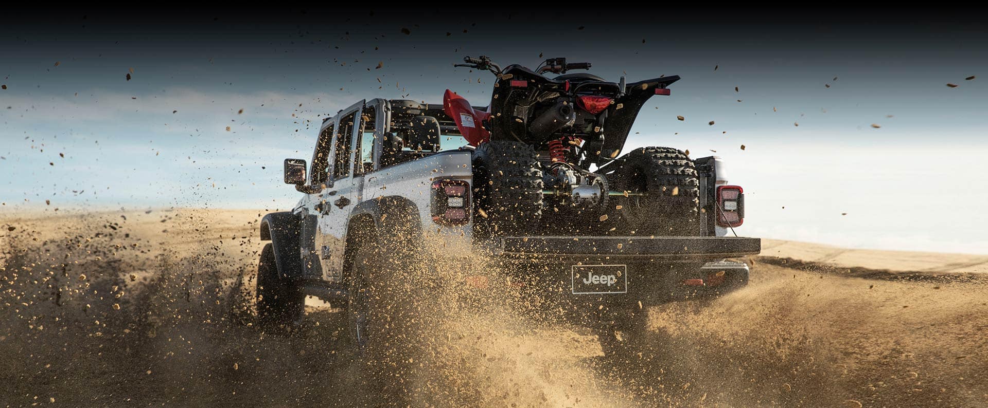 The rear view of a 2023 Jeep Gladiator Rubicon kicking up sand as it is driven across a desert, carrying an ATV in its bed with the tailgate open.
