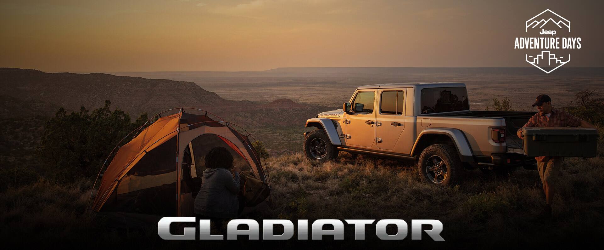  A driver-side rear profile of a 2023 Jeep Gladiator Rubicon parked on a hilltop at dusk with its tailgate open and a man carrying a cooler to a nearby campsite. Jeep Adventure Days logo. Gladiator.