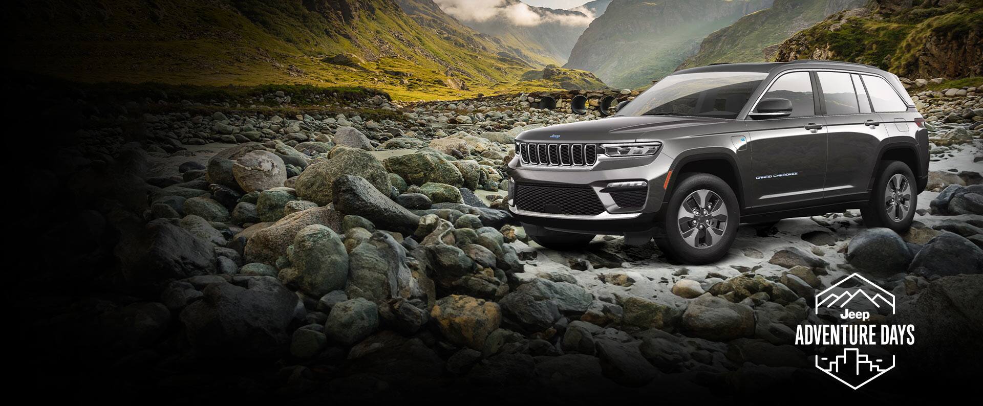 An angled driver-side profile of a gray 2023 Jeep Grand Cherokee 4xe parked on a bed of rocks off-road, with mountains in the background. Jeep Adventure Days logo.