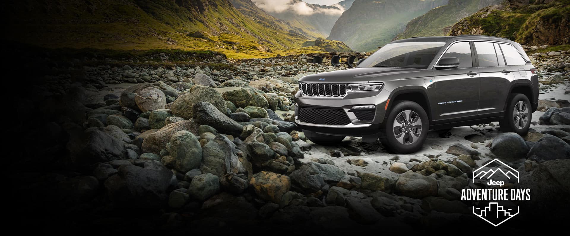 Un perfil desde el ángulo del conductor de un Jeep Grand Cherokee 4xe 2023 gris estacionado sobre un lecho de rocas fuera de la carretera, con montañas de fondo. Logo de Días de Aventura Jeep.
