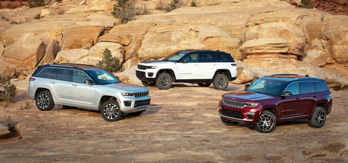 Display Three 2023 Jeep Grand Cherokee models, including the Grand Cherokee Overland, Summit Reserve and the Grand Cherokee 4xe, parked off-road on bare rock.