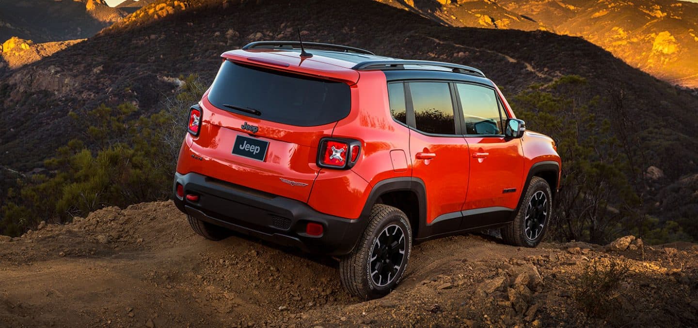 Display A rear three-quarter view of a red 2023 Jeep Renegade Trailhawk parked at the top of an off-road trail, with the sunset reflecting across mountains in the background. 