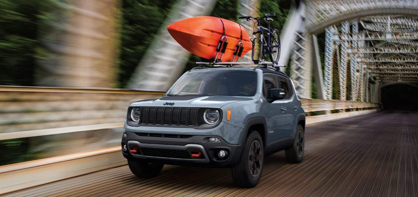 Display A gray 2023 Jeep Renegade Trailhawk with a kayak and bicycle strapped to its roof rack, being driven across a bridge with the background blurred to indicate the speed of the vehicle.