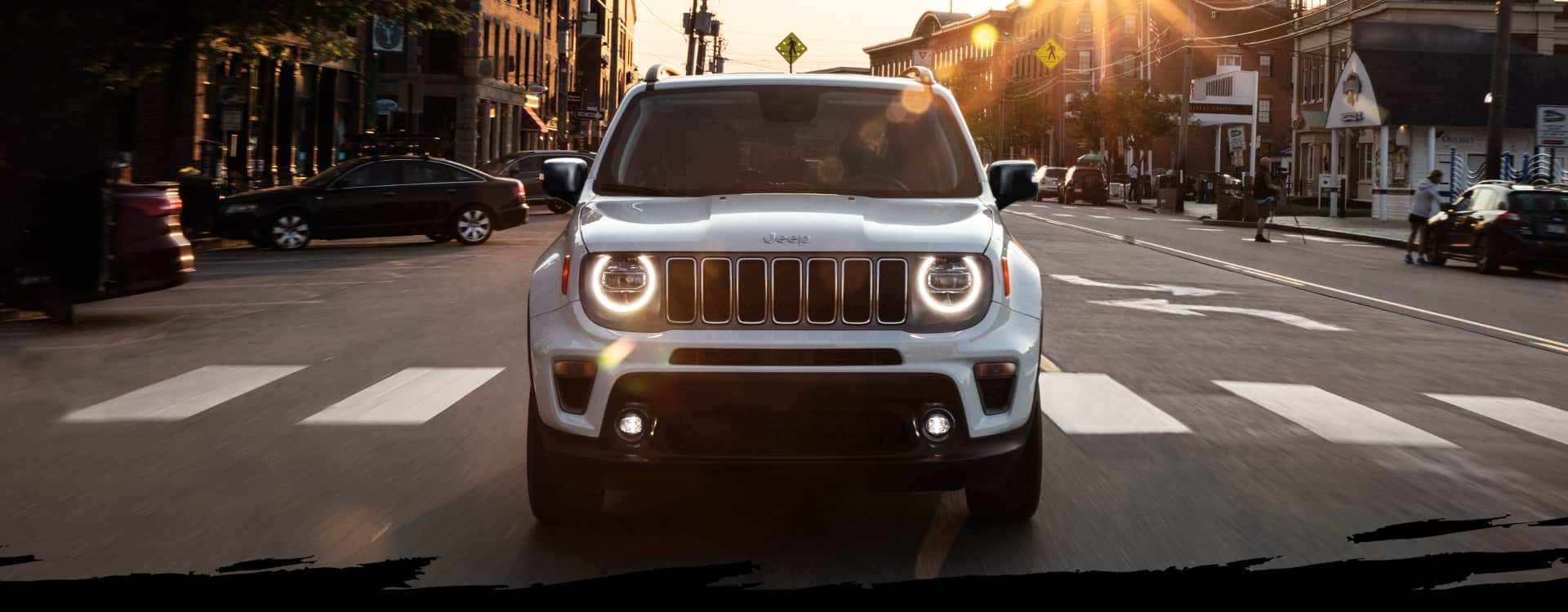 A head-on view of the 2023 Jeep Renegade Limited being driven on a busy city street at sunset, with its headlamps on.