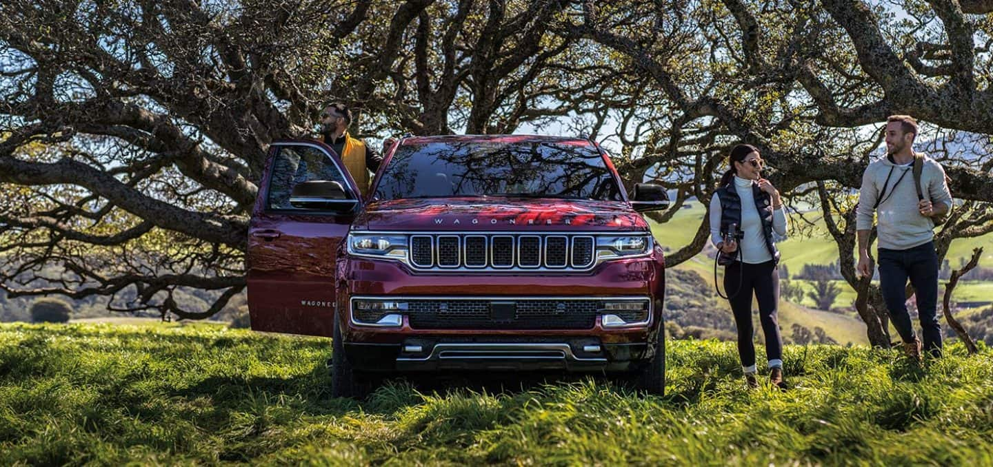 Display A group of friends with rucksacks and a camera standing beside a 2023 Wagoneer Series III that is parked off-road under s sprawling tree canopy.