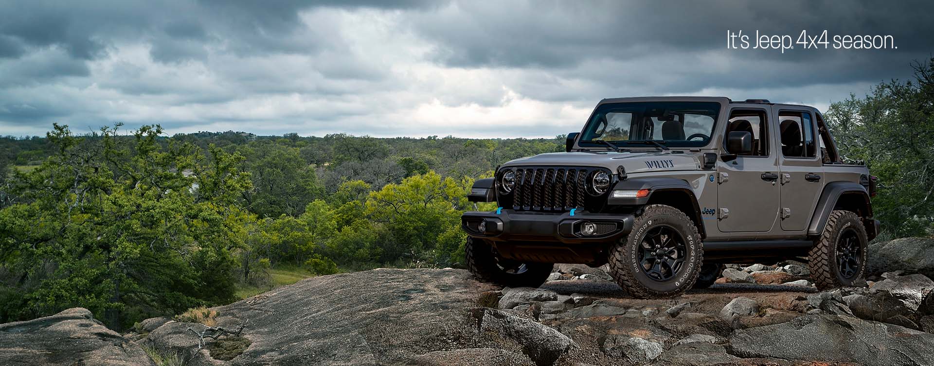 A gray 2023 Jeep Wrangler Willys 4xe parked on a rocky outcropping with a stormy sky above and a forest of trees below. The It's Jeep Season logo.