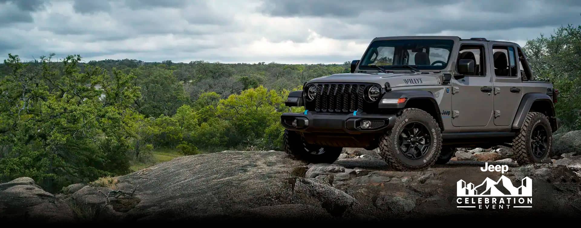 A gray 2023 Jeep Wrangler Willys 4xe parked on a rocky outcropping with a stormy sky above and a forest of trees below. The Jeep Celebration Event.