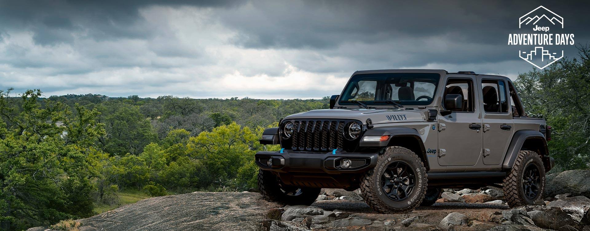 A gray 2023 Jeep Wrangler Willys 4xe parked on a rocky outcropping with a stormy sky above and a forest of trees below. Jeep Adventure Days logo.