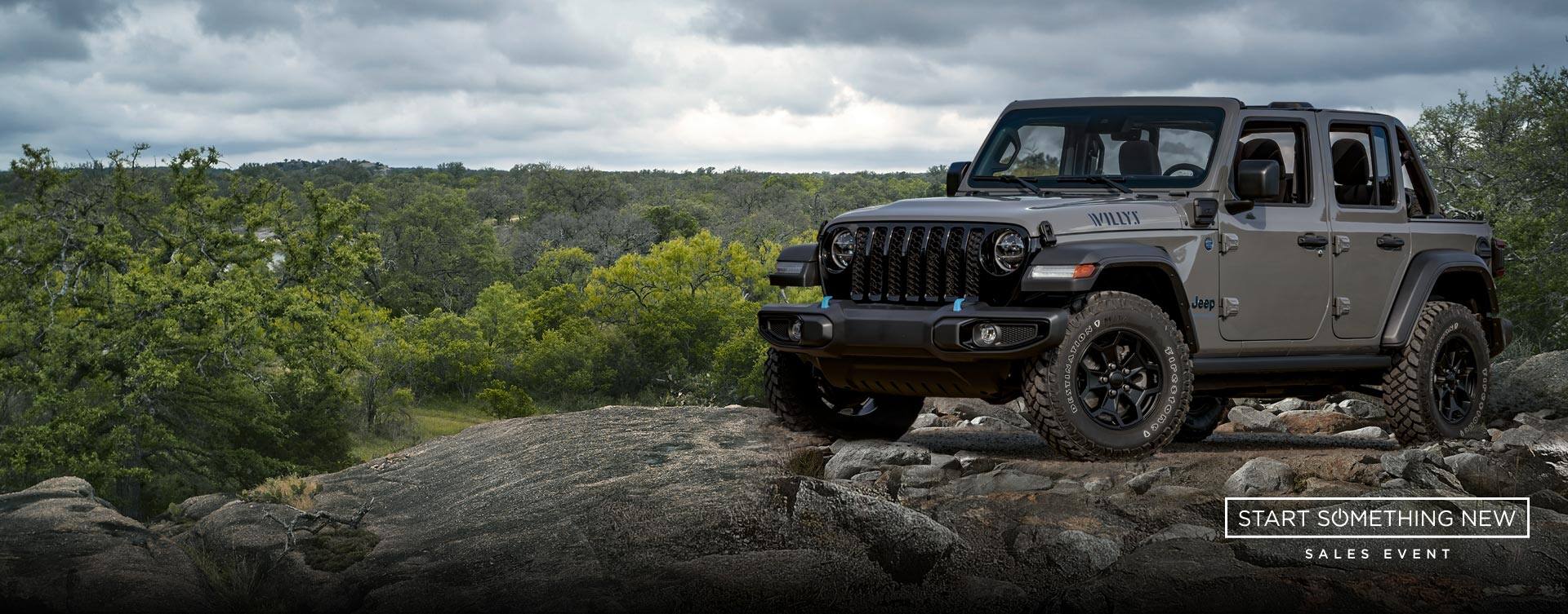 A gray 2023 Jeep Wrangler Willys 4xe parked on a rocky outcropping with a stormy sky above and a forest of trees below. The start something new sales event logo.