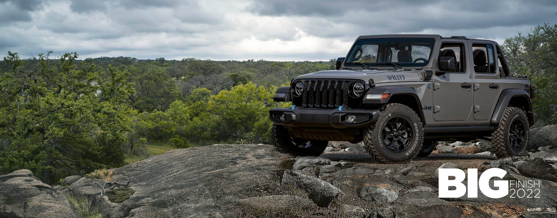 A gray 2023 Jeep Wrangler Willys 4xe parked on a rocky outcropping with a stormy sky above and a forest of trees below. The Big Finish 2022 logo.