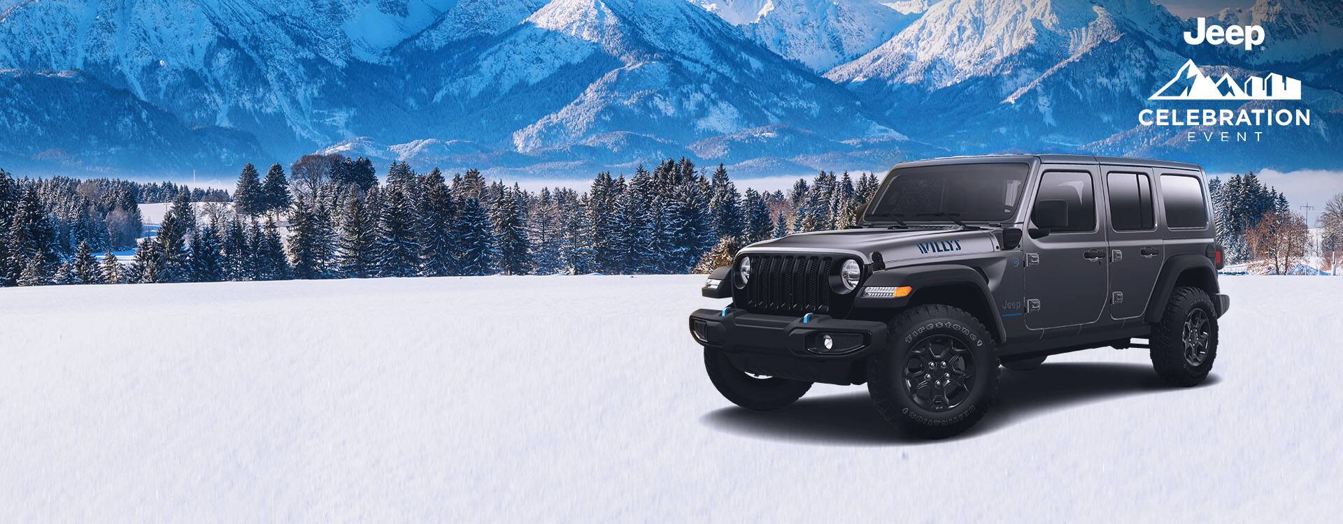An angled driver-side profile of a gray 2023 Jeep Wrangler Willys 4xe parked on snow, with evergreen trees and mountains in the background. Jeep. Jeep Celebration Event logo.
