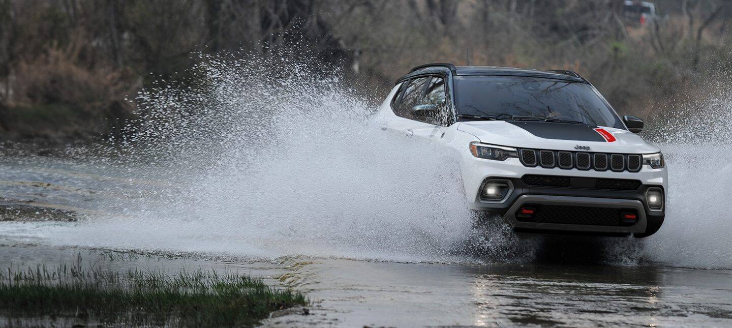 A 2024 Jeep Compass Trailhawk being driven through a stream with water splashing up the sides of the doors.