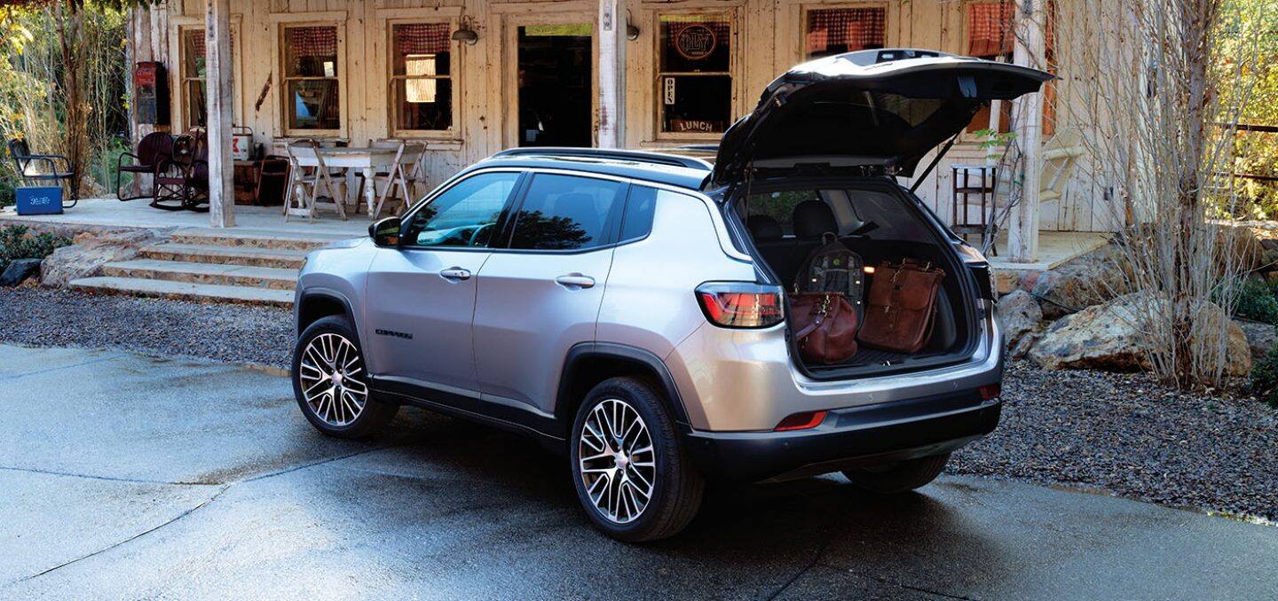 Display A 2024 Jeep Compass Limited parked at an old country store. Its liftgate is open revealing several pieces of luggage in the cargo area.