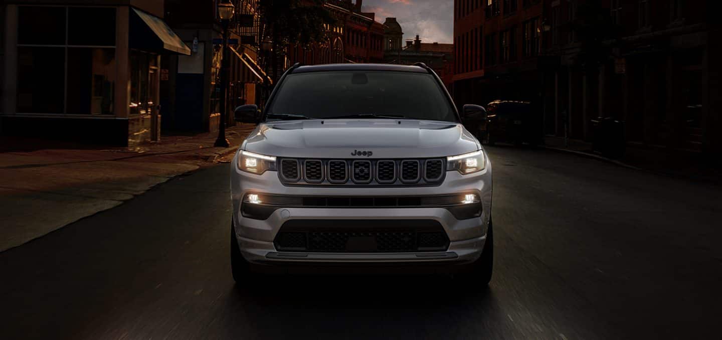 Display A head-on angle of a silver 2024 Jeep Compass High Altitude being driven on a city street surrounded by a variety of shops.