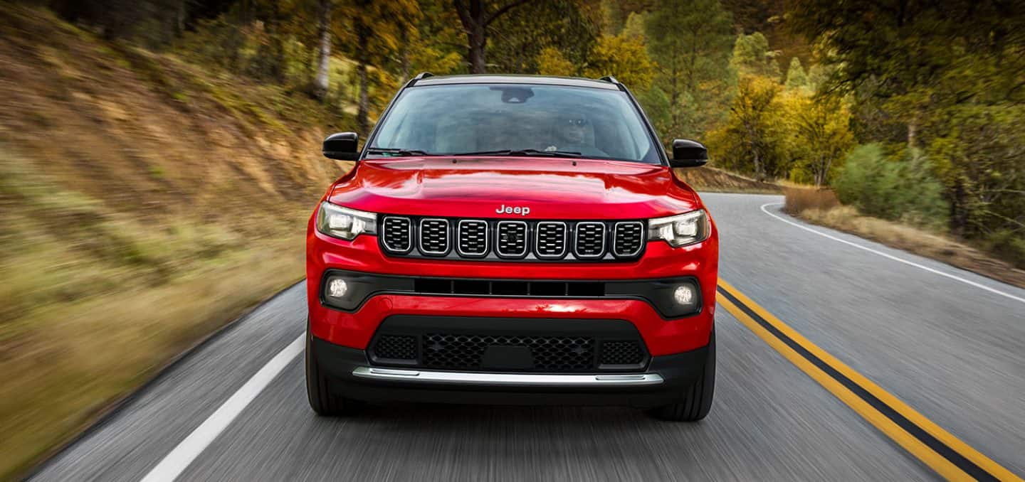 Display A head-on angle of a red 2024 Jeep Compass High Altitude being driven on a winding highway in the woods.