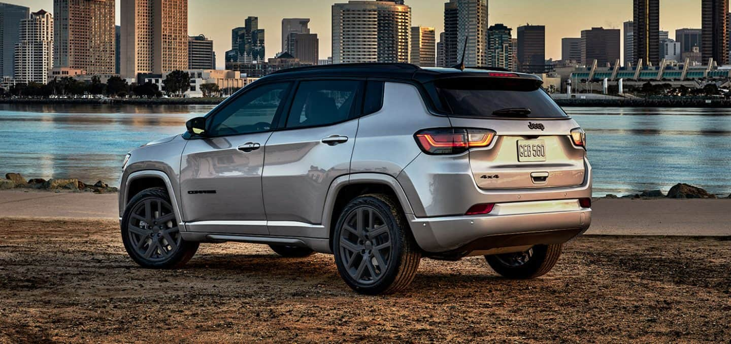 Display A rear driver-side profile of a silver 2024 Jeep Compass High Altitude parked beside the riverfront with tall buildings across the water.