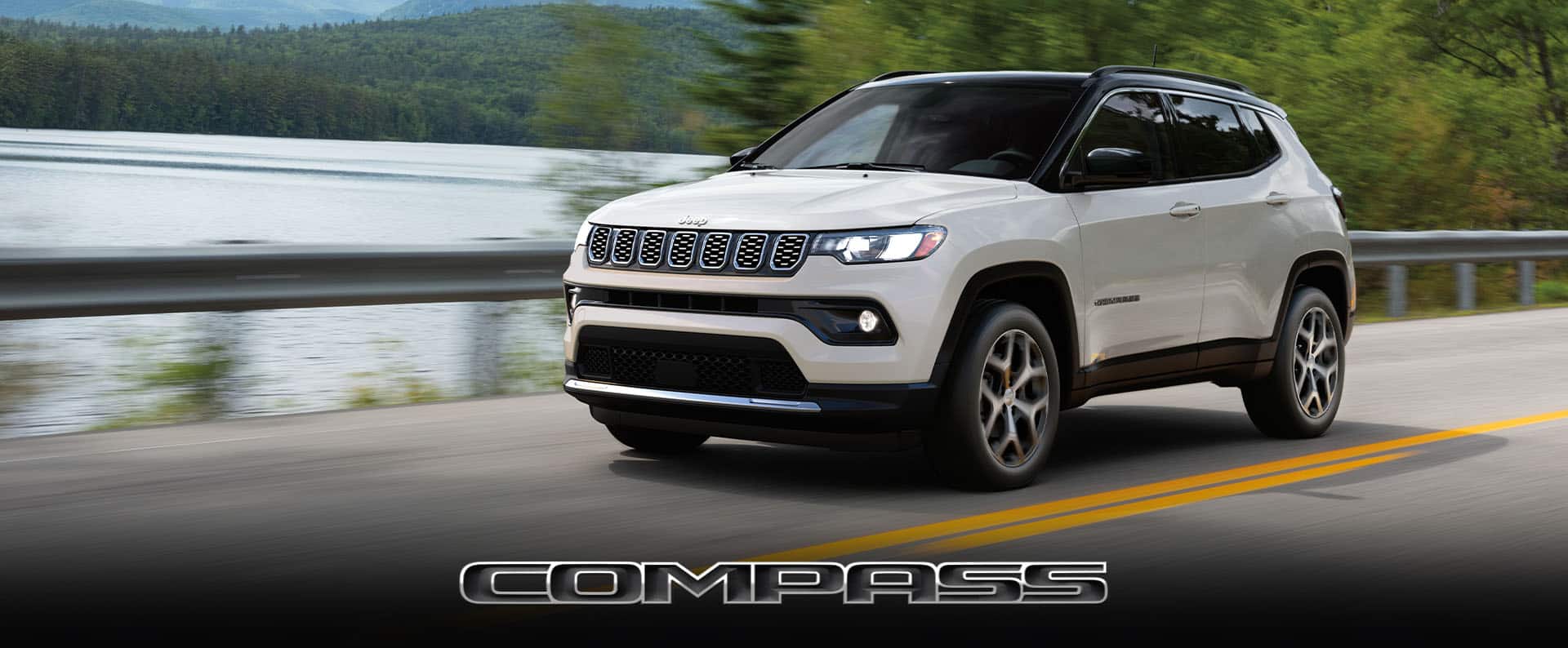 A white 2024 Jeep Compass Limited being driven on a highway beside a lake.