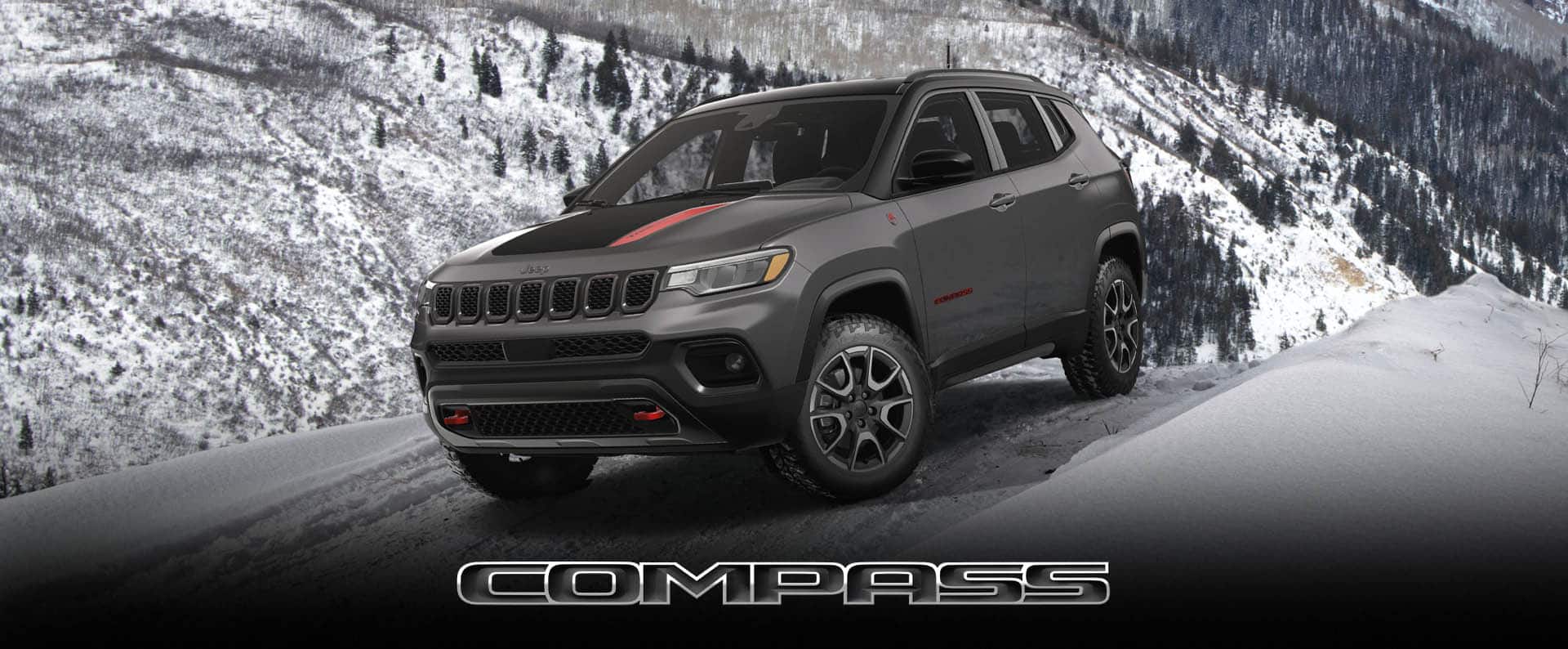 A gray 2024 Jeep Compass Trailhawk with a black and red hood insert being driven down a snow-covered hill in the mountains. Compass.