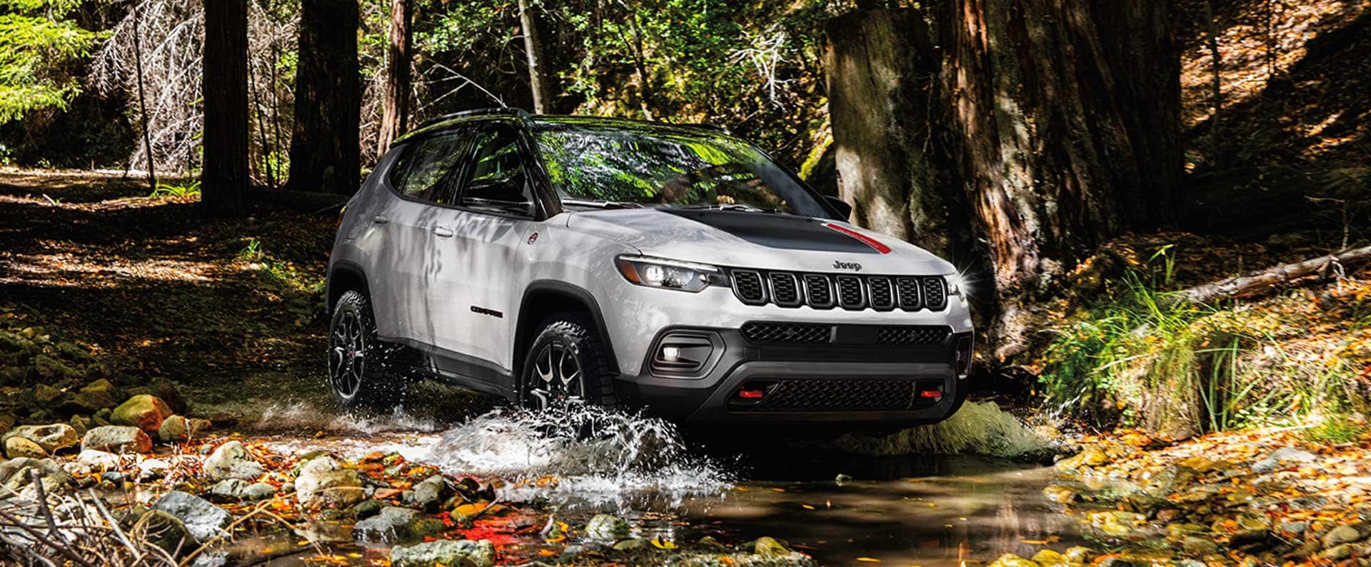 A white 2024 Jeep Compass Trailhawk with a black hood insert, fording through a shallow stream off-road in the woods. Employee Pricing Plus event. 