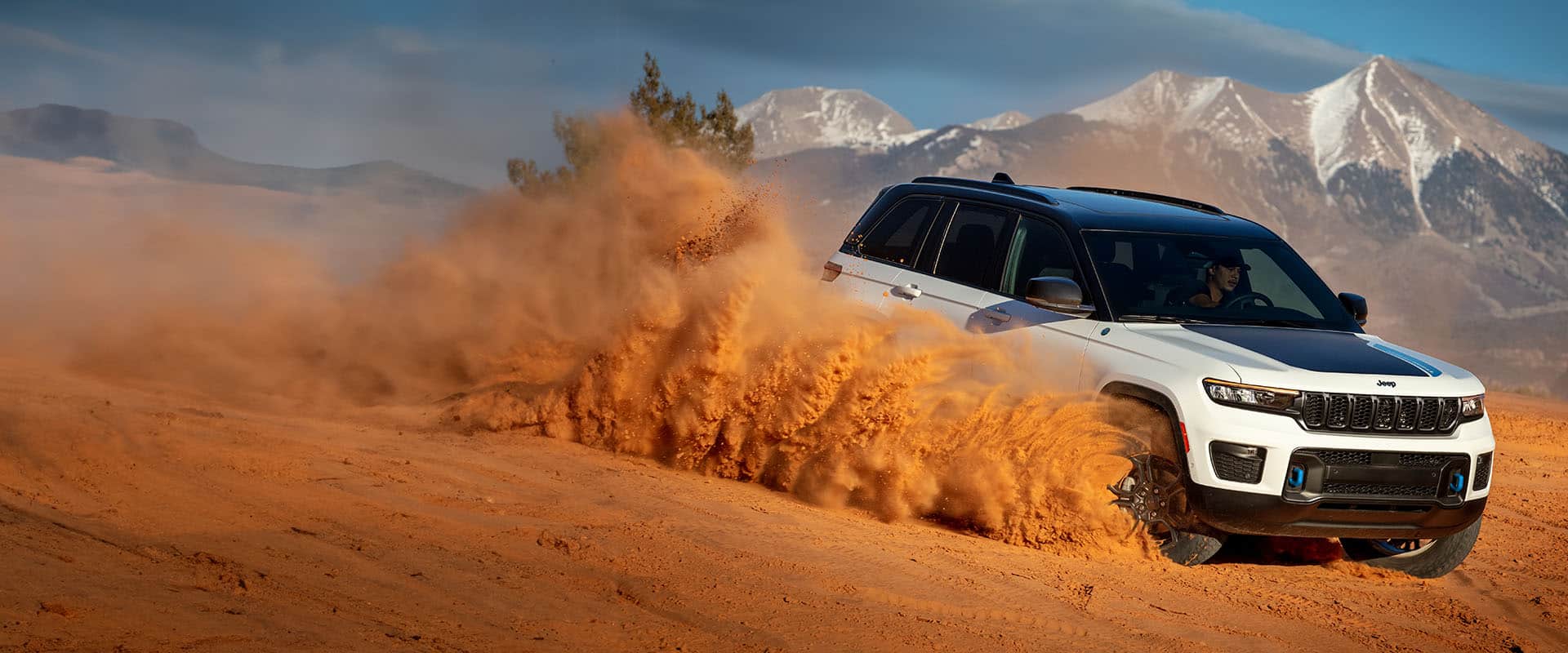 A white 2024 Jeep Grand Cherokee 4xe Trailhawk kicking up a cloud of orange sand and dust as it's being driven down a dune, with mountains in the background. 